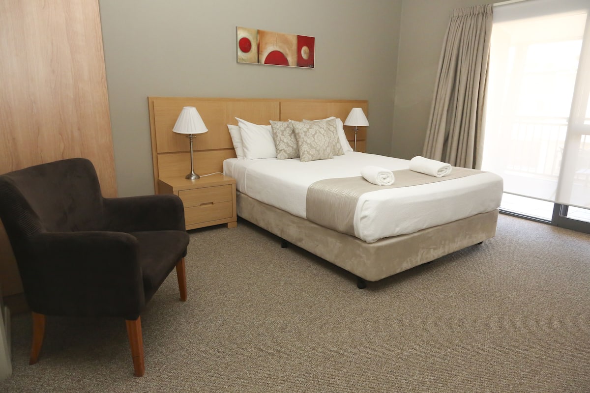 Una habitación de hotel con una cama, dos lámparas de noche, una silla y un cuadro en la pared.