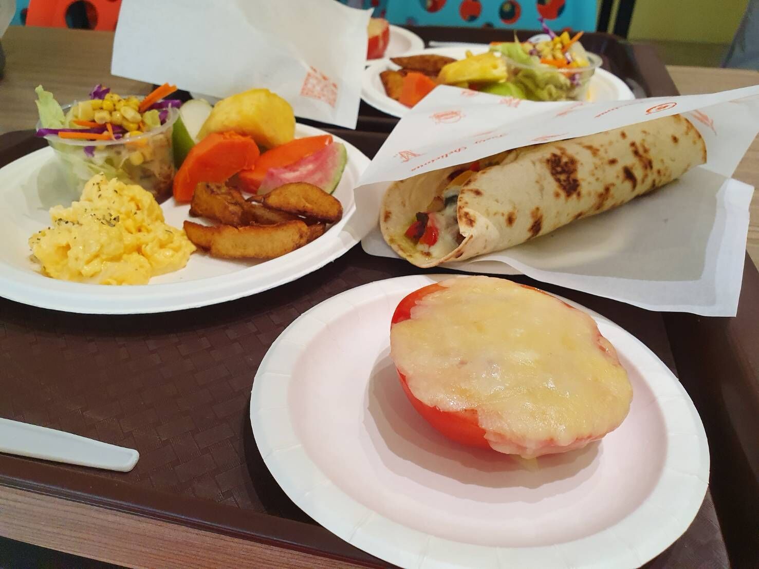 A tray with a plate of scrambled eggs, fried potatoes, a salad, a wrap, and a slice of tomato topped with cheese.