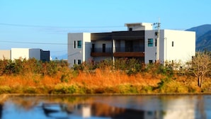 A modern two-story building with a flat roof and large windows, surrounded by natural vegetation and a body of water.