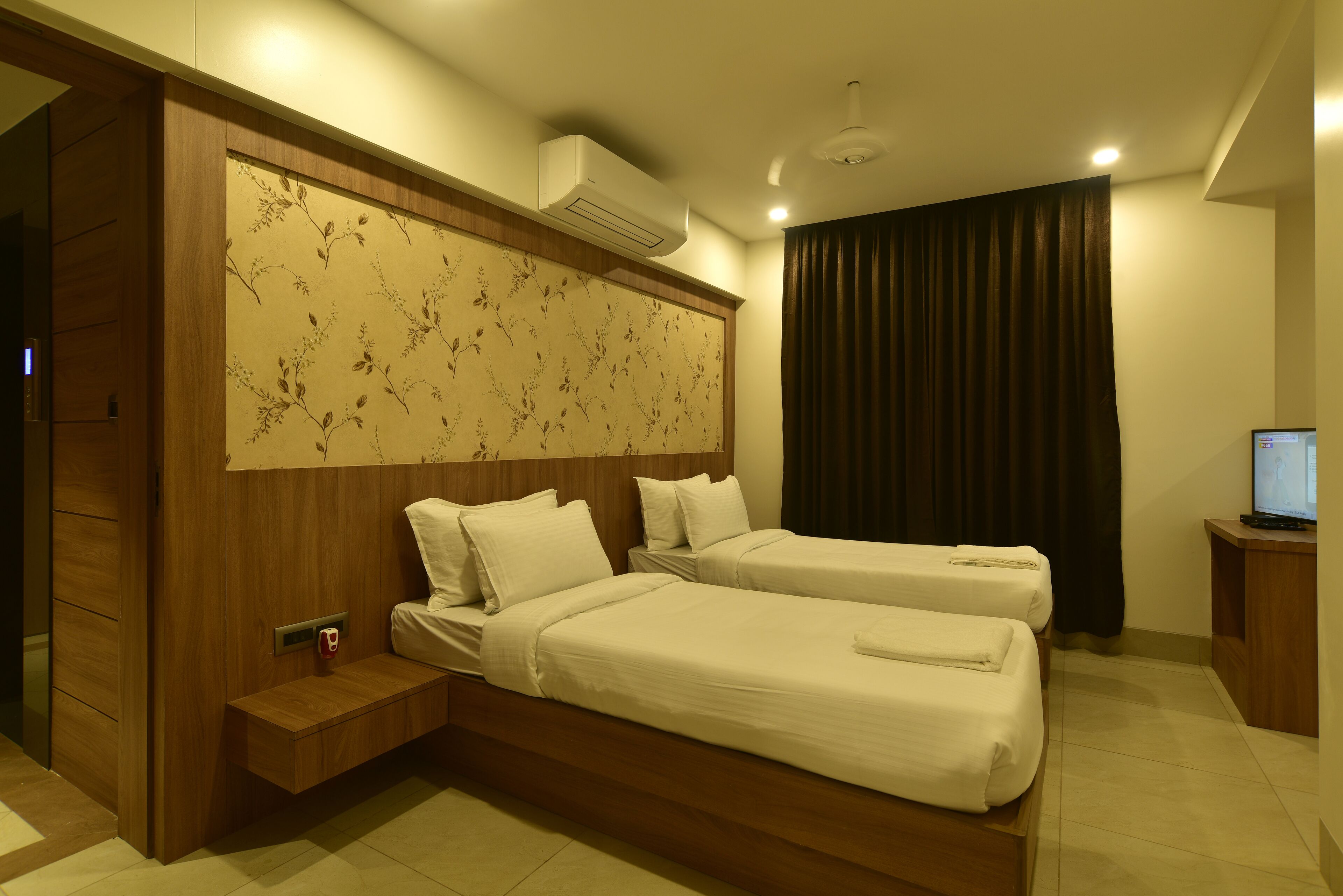 A hotel room with two beds, a wooden headboard, a wall-mounted air conditioner, a small bedside table, and a television on a wooden stand.