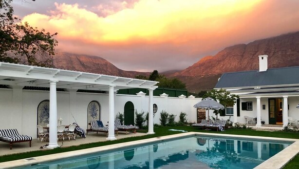 A swimming pool with lounge chairs and umbrellas, surrounded by a white pergola and a house with a mountain backdrop. - La Cotte The Manor House (Franschhoek)