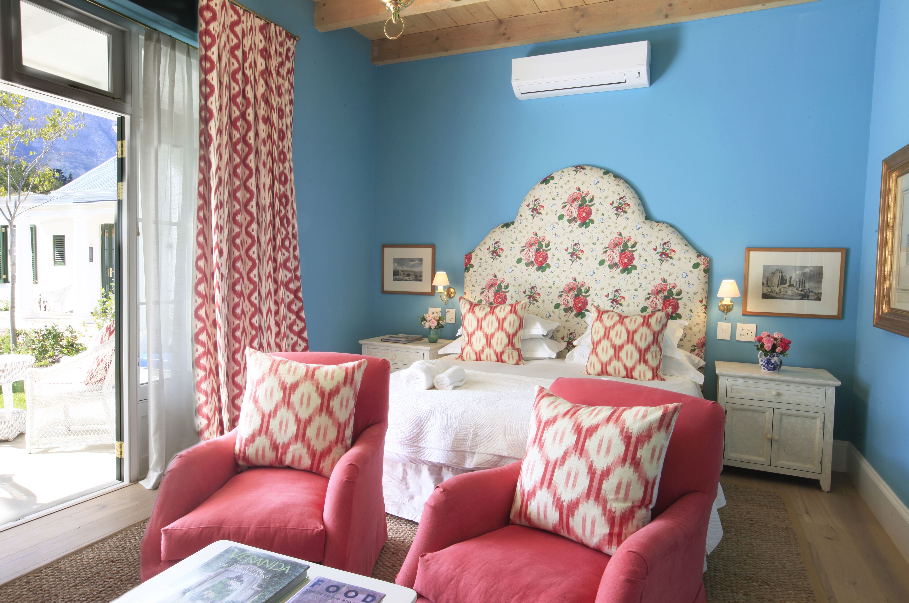 Un dormitorio con pared azul, cabecera floral, dos sillones rojos, cama blanca y ventana con vista a un edificio y follaje.