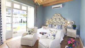 A bedroom with a large bed, two white armchairs, a wooden ceiling, and a view of a swimming pool. - La Cotte The Manor House (Franschhoek)