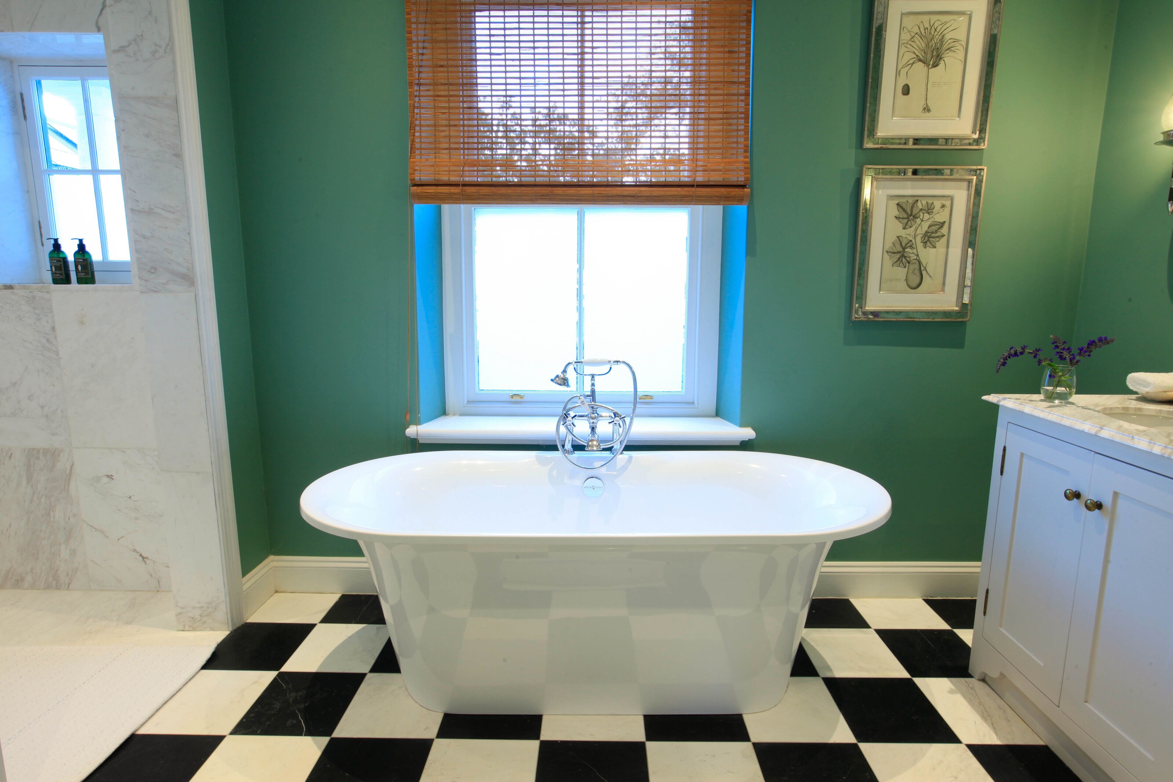 A bathroom with a freestanding bathtub, a window with blinds, a checkered floor, and framed botanical artwork on the wall.