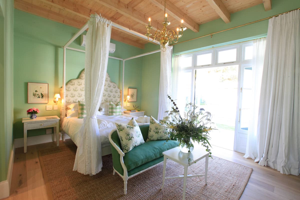 Ein Bett mit Baldachin, ein Nachttisch mit Lampe, ein grünes Sofa mit floralen Kissen und eine Vase mit Blumen.