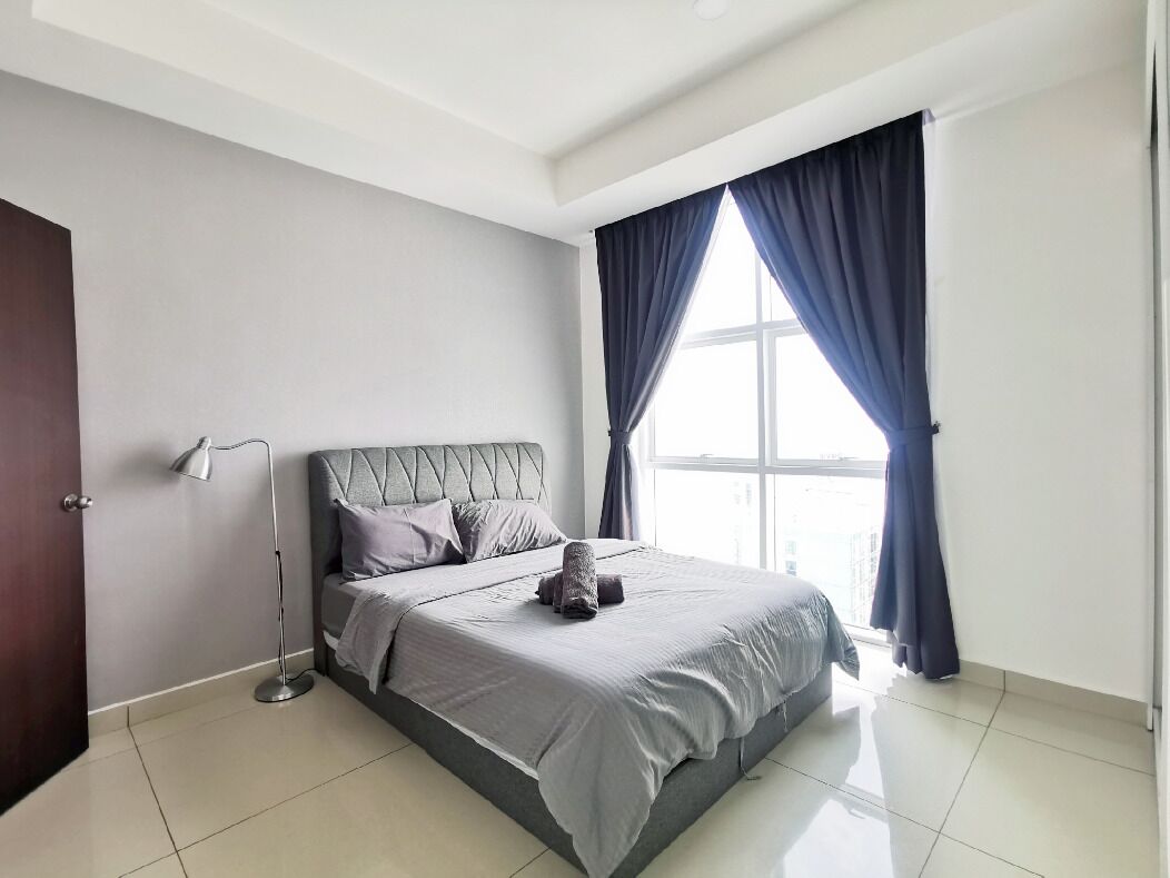 A modern bedroom with a large bed, a bedside lamp, and a view of the city through a window with curtains.