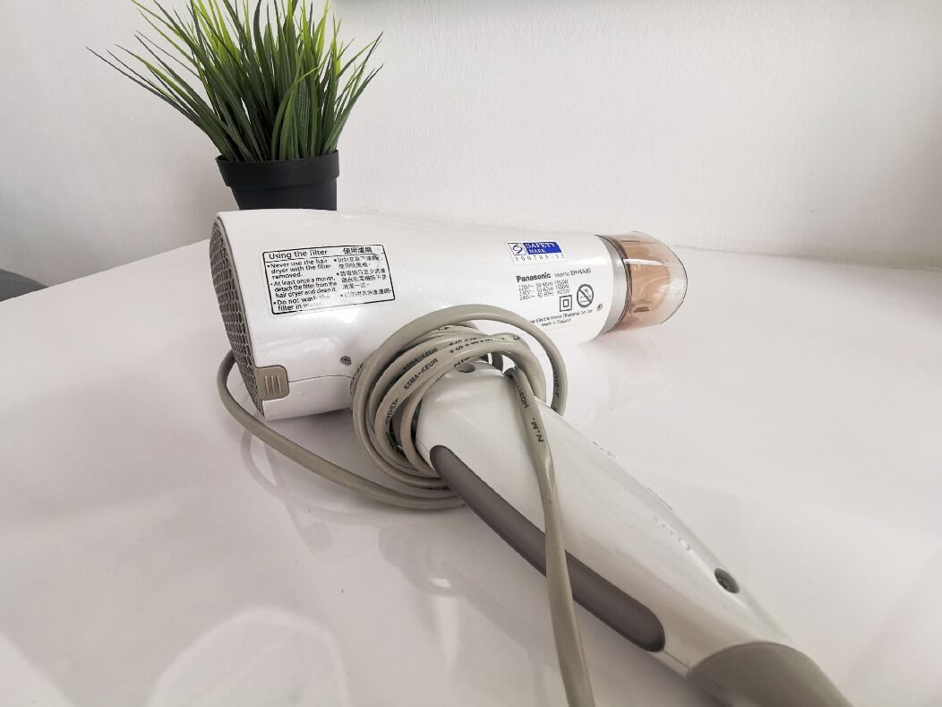 Een witte haardroger met snoer en een etiket, geplaatst op een witte ondergrond naast een bloempot.