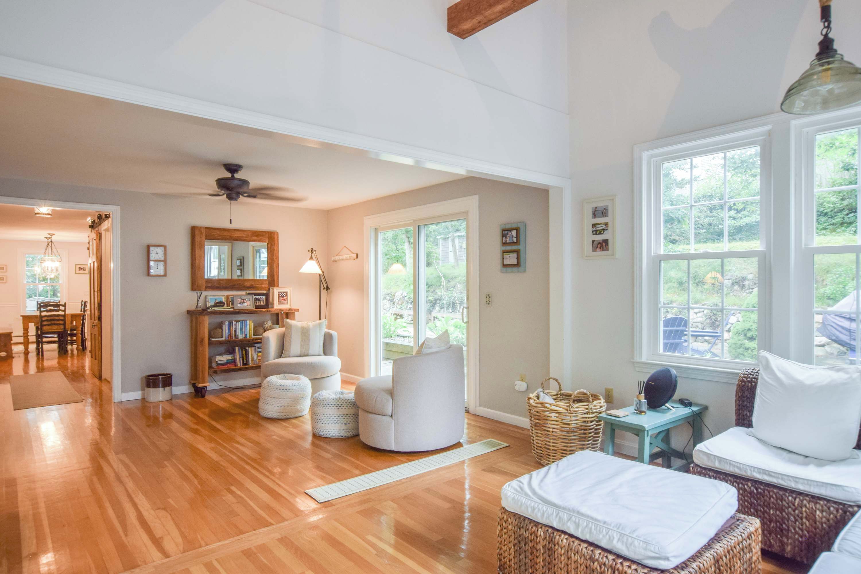 A spacious living room with hardwood flooring, a ceiling fan, a large window with a view of greenery, and a cozy seating area with a sofa and ottoman.