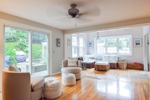 A spacious living room with a large window, a ceiling fan, and a seating area with ottomans and a sofa.