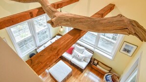 A loft-style room with exposed wooden beams, a large window, and a wicker sofa with cushions. - Spacious w/ Large Outdoor Area Fire Pit! (Chatham)