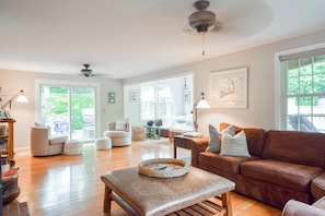 A spacious living room with a brown leather couch, a wooden coffee table, and a view of a garden through large windows.