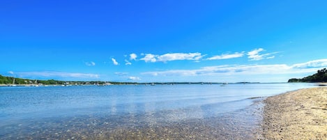 A beach with clear water, pebbles, and boats in the distance.