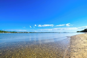 A beach with clear water, pebbles, and boats in the distance.