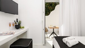 A modern hotel room with a flat-screen TV, a small table with a chair, a bed with rolled towels, a window with curtains, and a potted plant. - Two Bedroom Apartment, Diadema Apartments Naxos (Naxos)