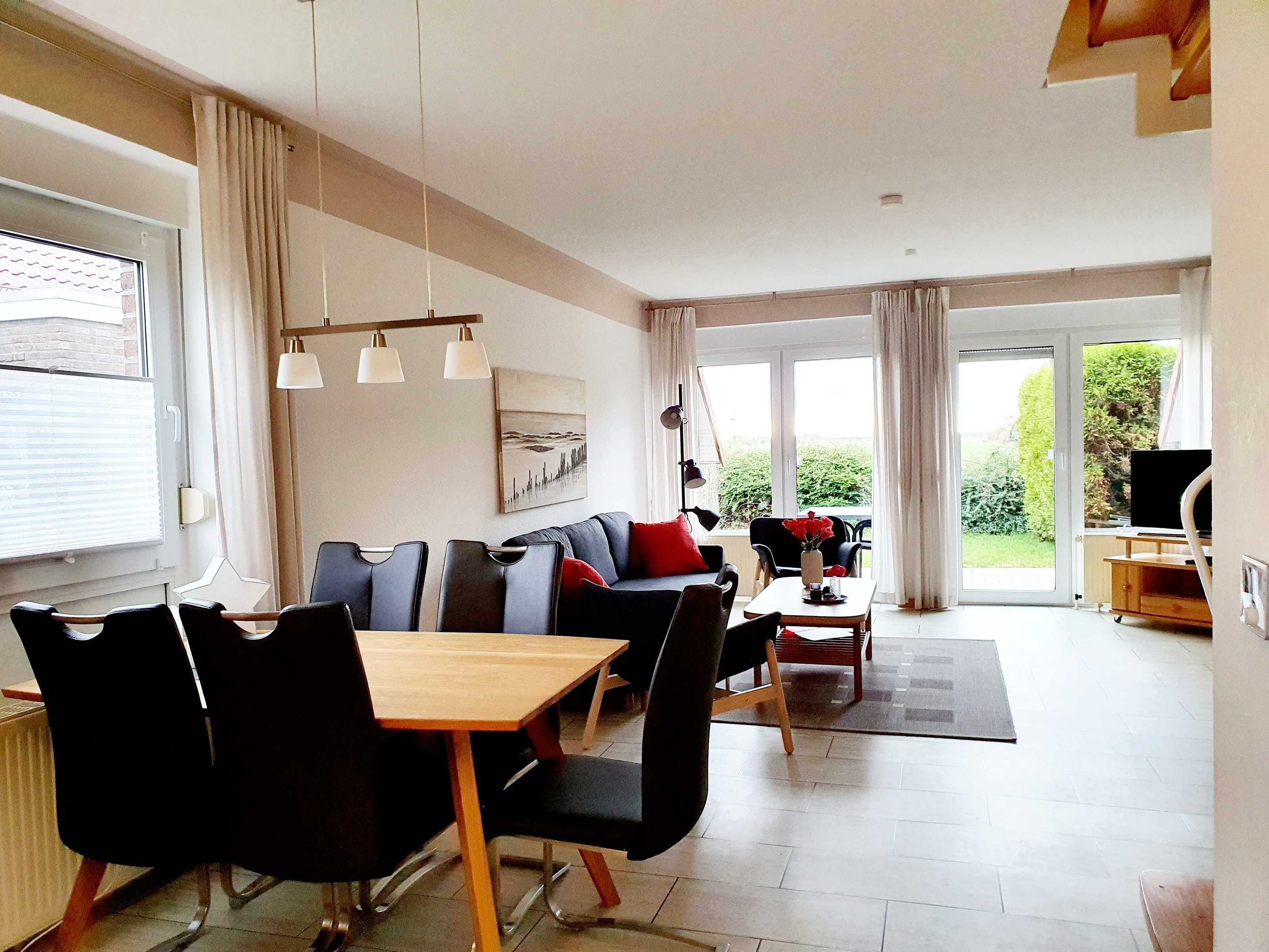 A modern living room with a sofa, dining table, and chairs, large windows with curtains, and a flat-screen TV.