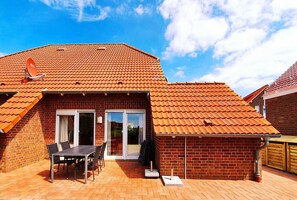Une maison en briques avec un toit en tuiles, une terrasse avec une table et des chaises, et une porte-fenêtre coulissante donnant sur un espace abrité.