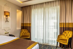 Hotellihuone, jossa on suuri sänky, kaksi nojatuolia, lamppu ja näkymä kaupunkiin ohuiden verhojen läpi.