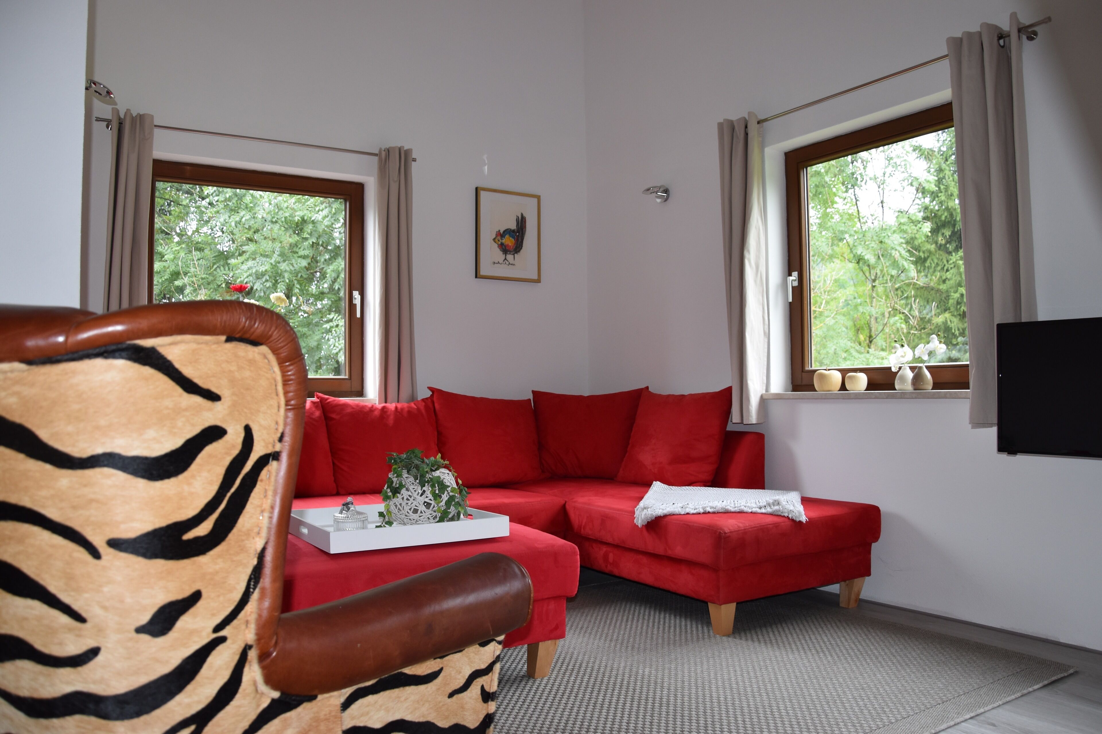 A living room with a red sofa, a tiger-patterned armchair, and a window with curtains.