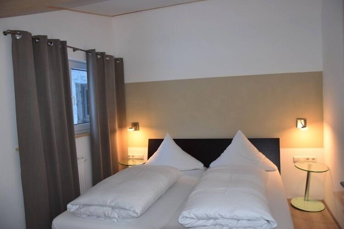 Una cama doble con ropa blanca, dos mesitas de noche con lámparas y una ventana con cortinas.