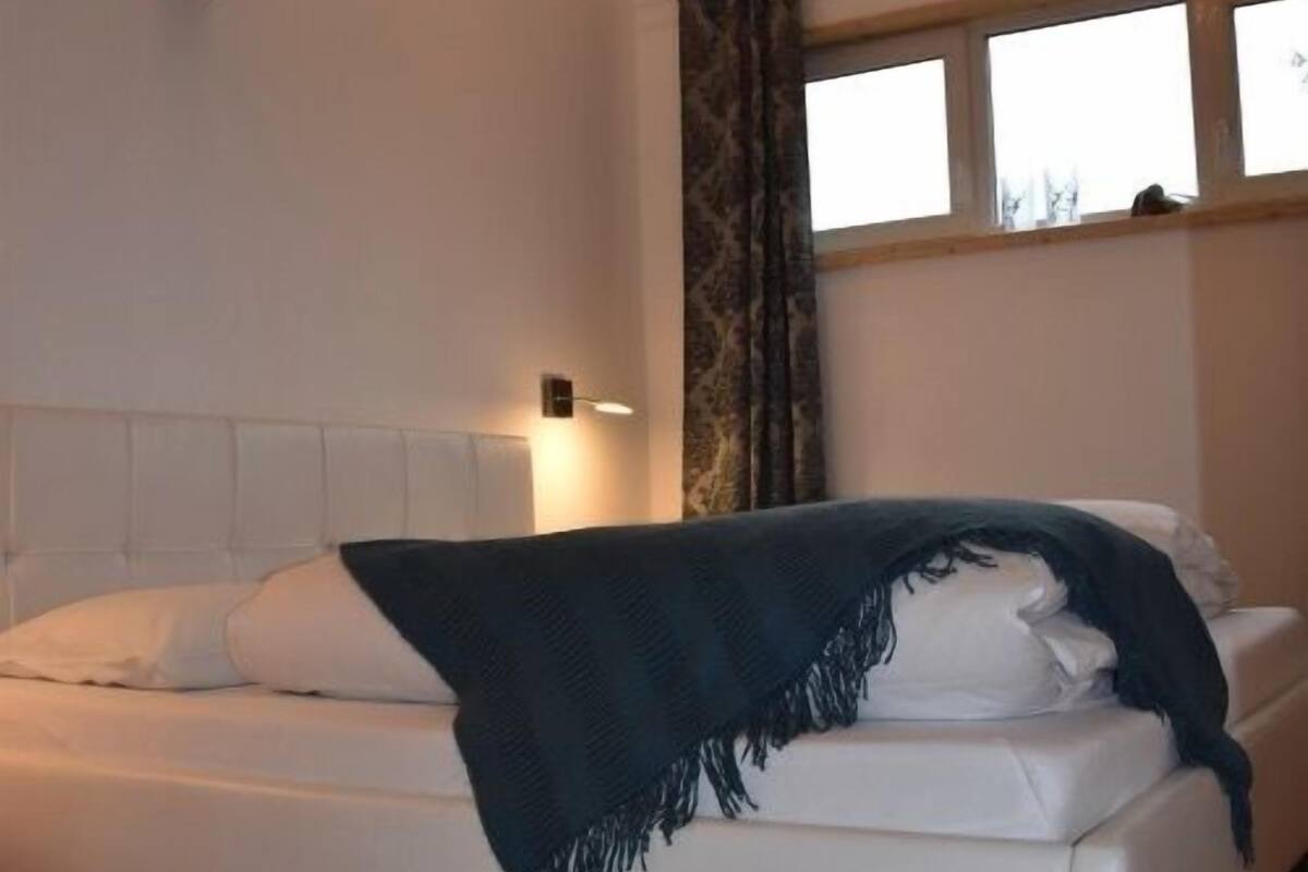 Un dormitorio con cama blanca, vigas de madera, una ventana con cortinas y una lámpara fijada a la pared.