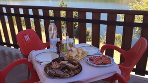 A table set with plates of food, a bottle, and condiments on a terrace overlooking a lake. - Camping Los Molinos (Palacios de Sanabria)