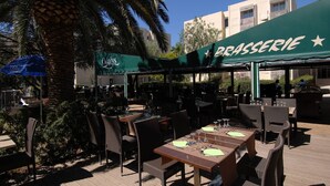 Buiten zitplaatsen met tafels en stoelen, een groen zonnescherm waarop 'BRASSERIE' staat, en een palmboom op de achtergrond.