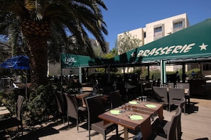 Sitzgelegenheiten im Freien mit Tischen und Stühlen, ein grüner Sonnenschirm mit der Aufschrift 'BRASSERIE' und eine Palme im Hintergrund.