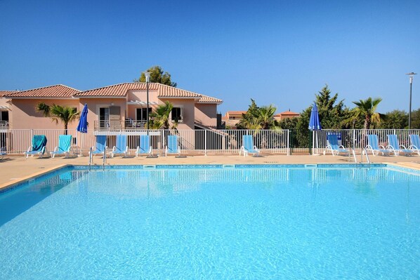 Une piscine entourée de chaises longues et de parasols, le tout délimité par une clôture, et un bâtiment avec un toit en tuiles en arrière-plan.