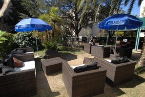 Terrasse mit Korbmöbeln und blauen Sonnenschirmen, umgeben von viel Grün.