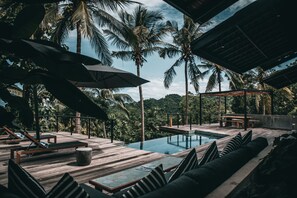 En træterrasse med liggestole og en pool, omgivet af palmer og udsigt til skoven.