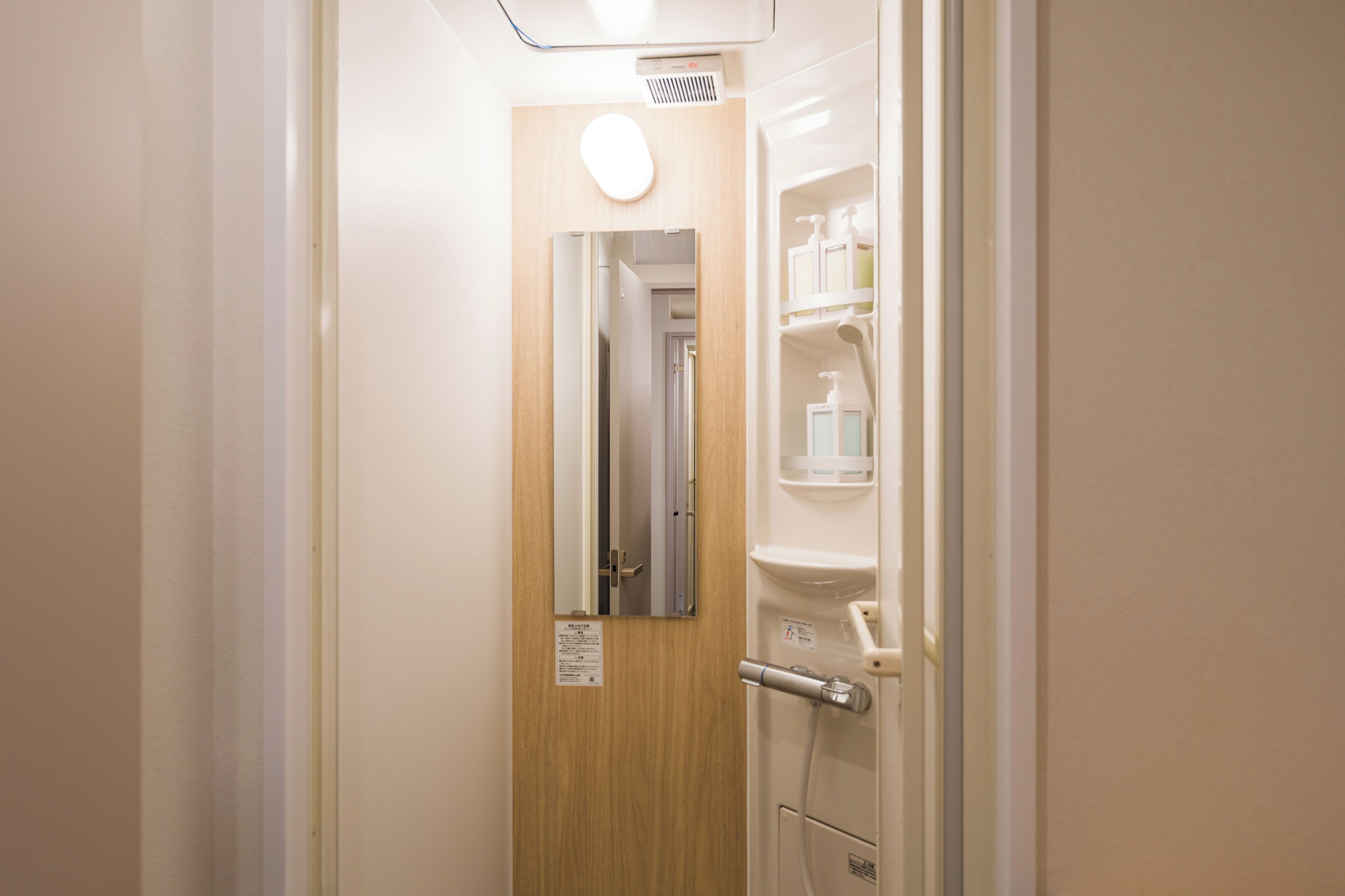 A hotel room with a wooden door, a mirror, and a white bathroom with a toilet, sink, and storage shelves.
