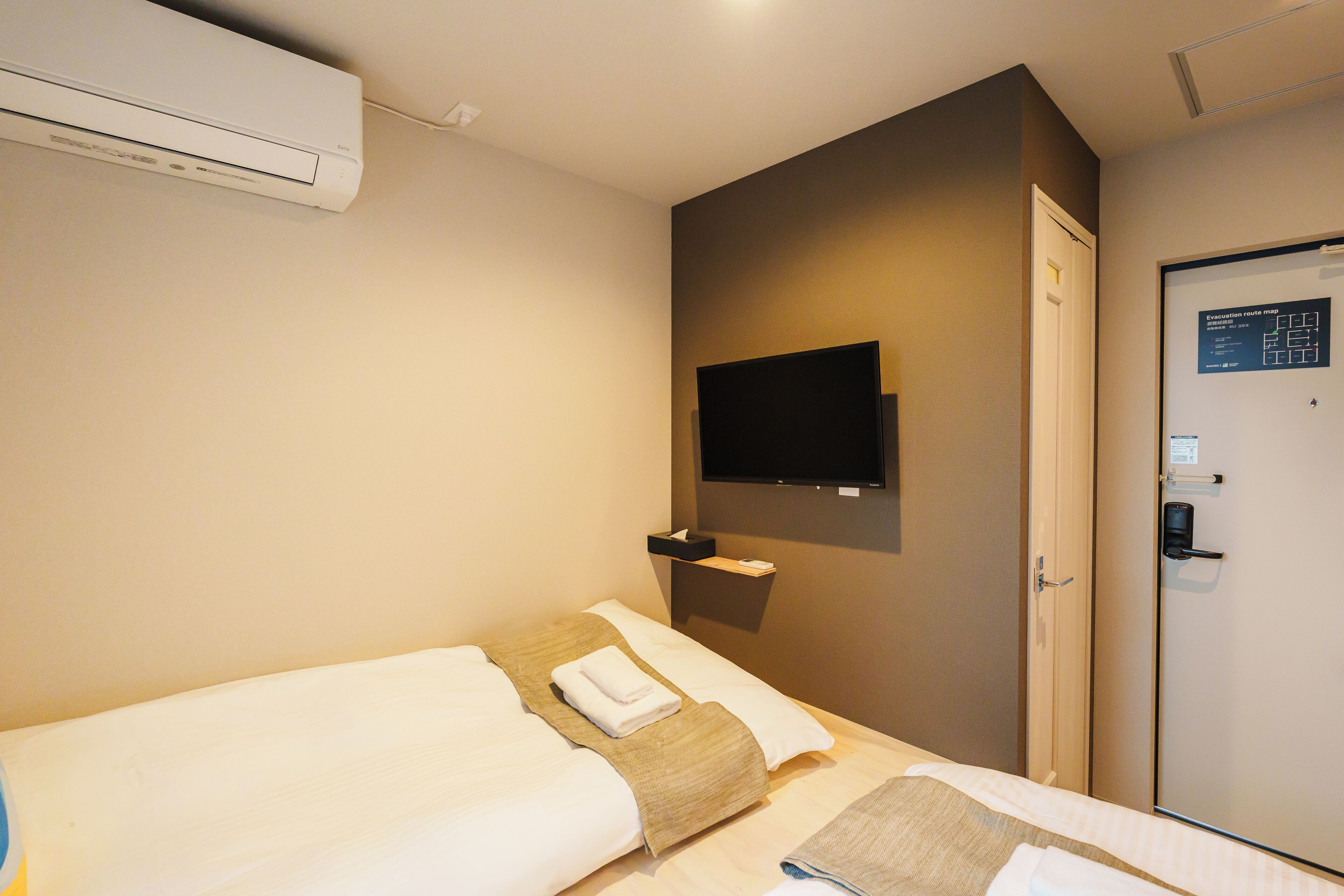A hotel room with two beds, a mounted TV, a door with a digital display, and an air conditioning unit.
