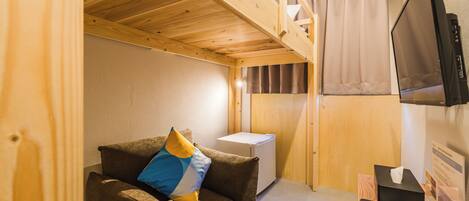Una habitación compacta con una cama litera, un pequeño sofá con un cojín, un televisor montado en la pared y un escritorio con un control remoto y una caja.