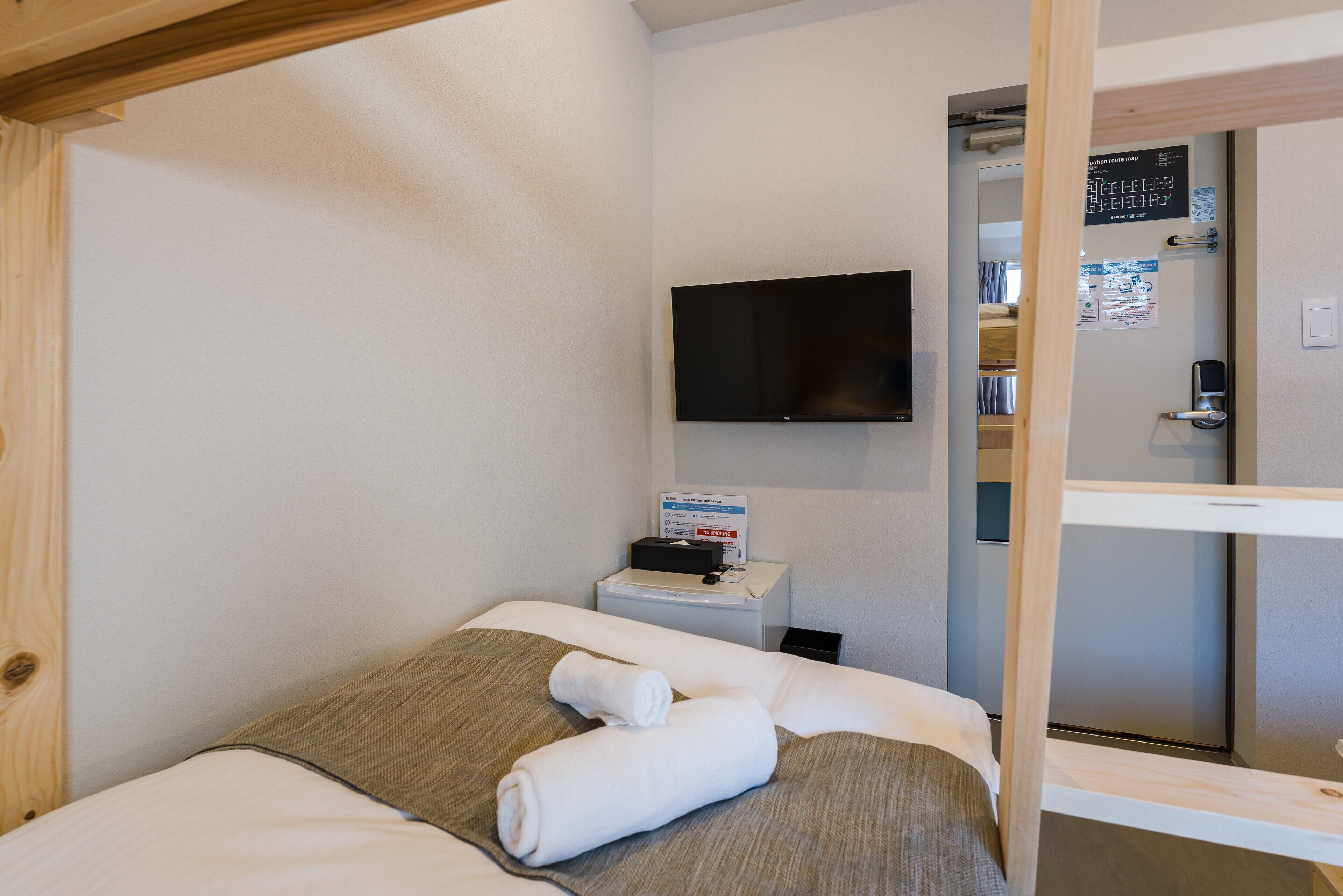 Habitación de hotel con cama, televisor de pantalla plana montado en la pared, mesita de noche con teléfono y una cajita, y ventana con marco de madera.