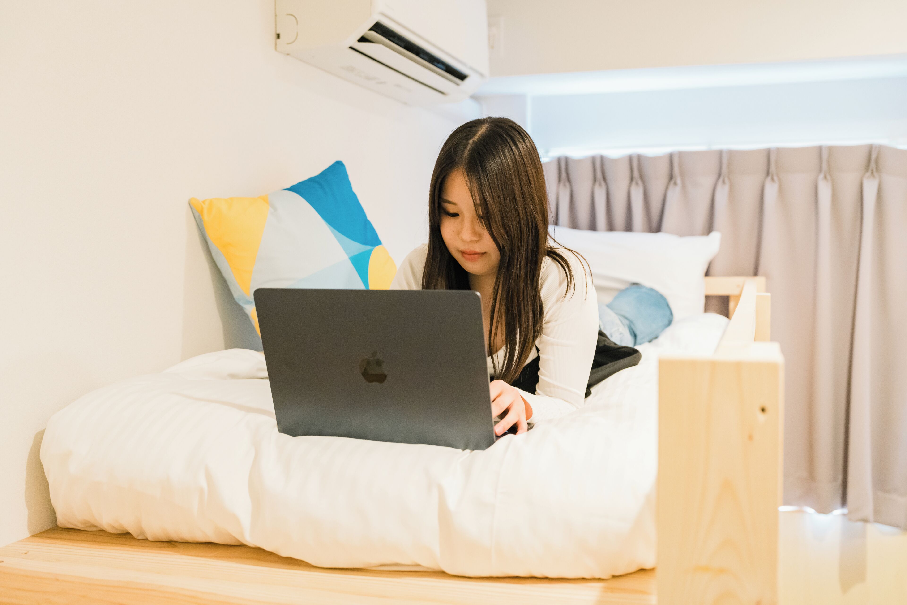A person using a laptop on a bed with a pillow, in a room with a wooden headboard and an air conditioning unit.