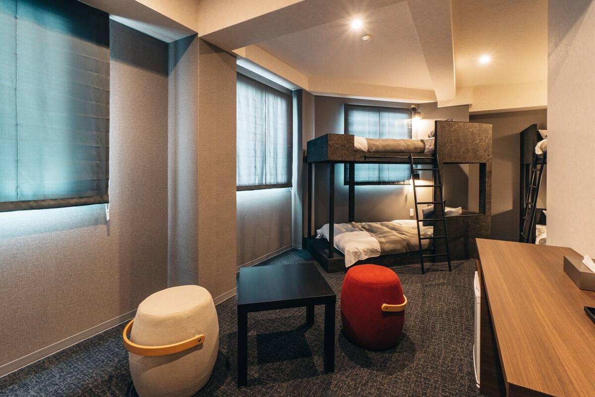 A compact hotel room with bunk beds, a small table, a red ottoman, and a desk.