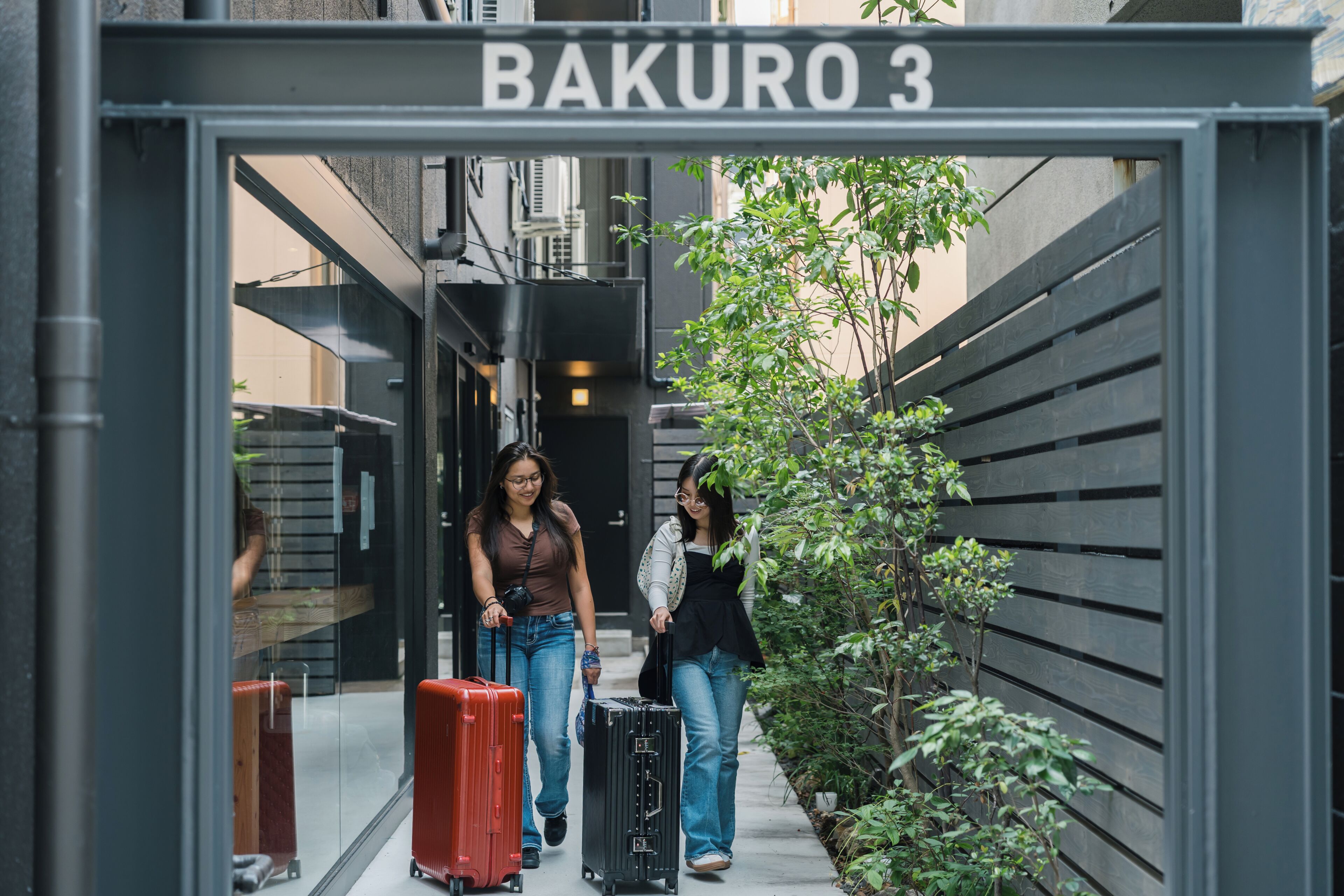 Une entrée menant à un bâtiment portant l’inscription « BAKURO 3 », avec un étroit chemin, un banc en bois et un petit arbre.