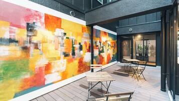 Une terrasse extérieure agrémentée d’une fresque murale abstraite aux couleurs vives, de tables et de chaises en bois, et d’une façade de bâtiment moderne.