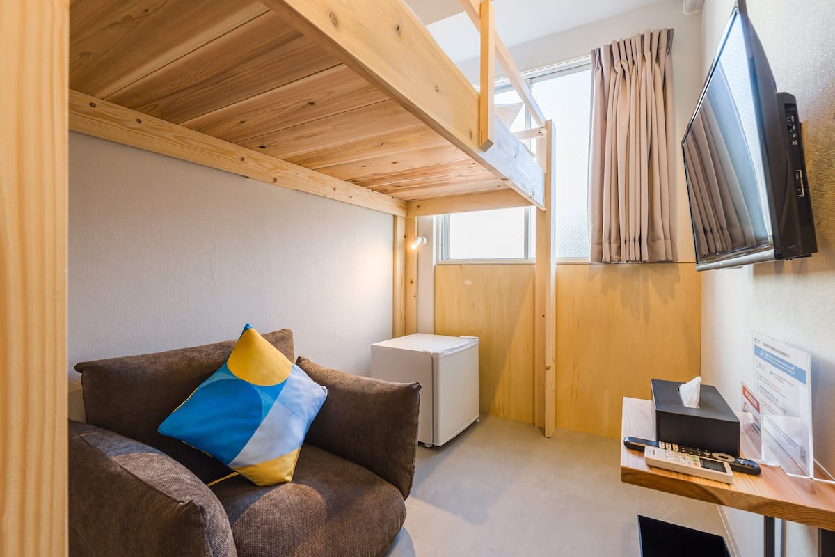 A cozy room with a wooden ceiling, a sofa with a blue and yellow pillow, a small white appliance, a wooden desk with a remote control, and a TV mounted on the wall.