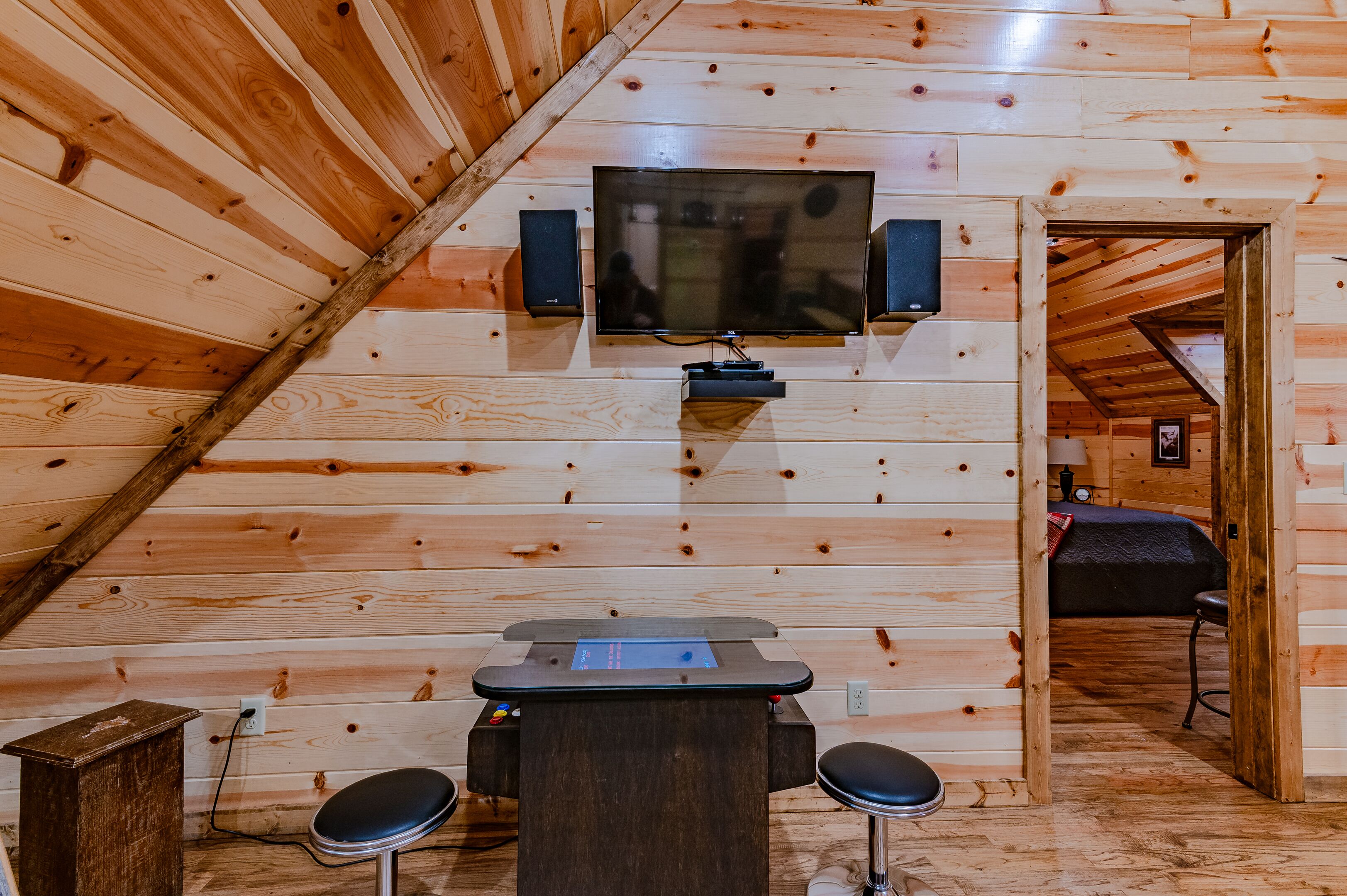 A cozy log cabin room with a wooden ceiling, a mounted television, and a small table with two stools.