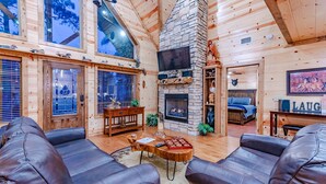 A cozy log cabin interior with a stone fireplace, leather sofas, and a large TV.