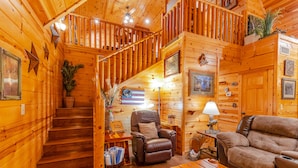 L’intérieur d’un chalet en bois, avec un escalier, des fauteuils inclinables en cuir, une lampe et des tableaux encadrés accrochés aux murs.