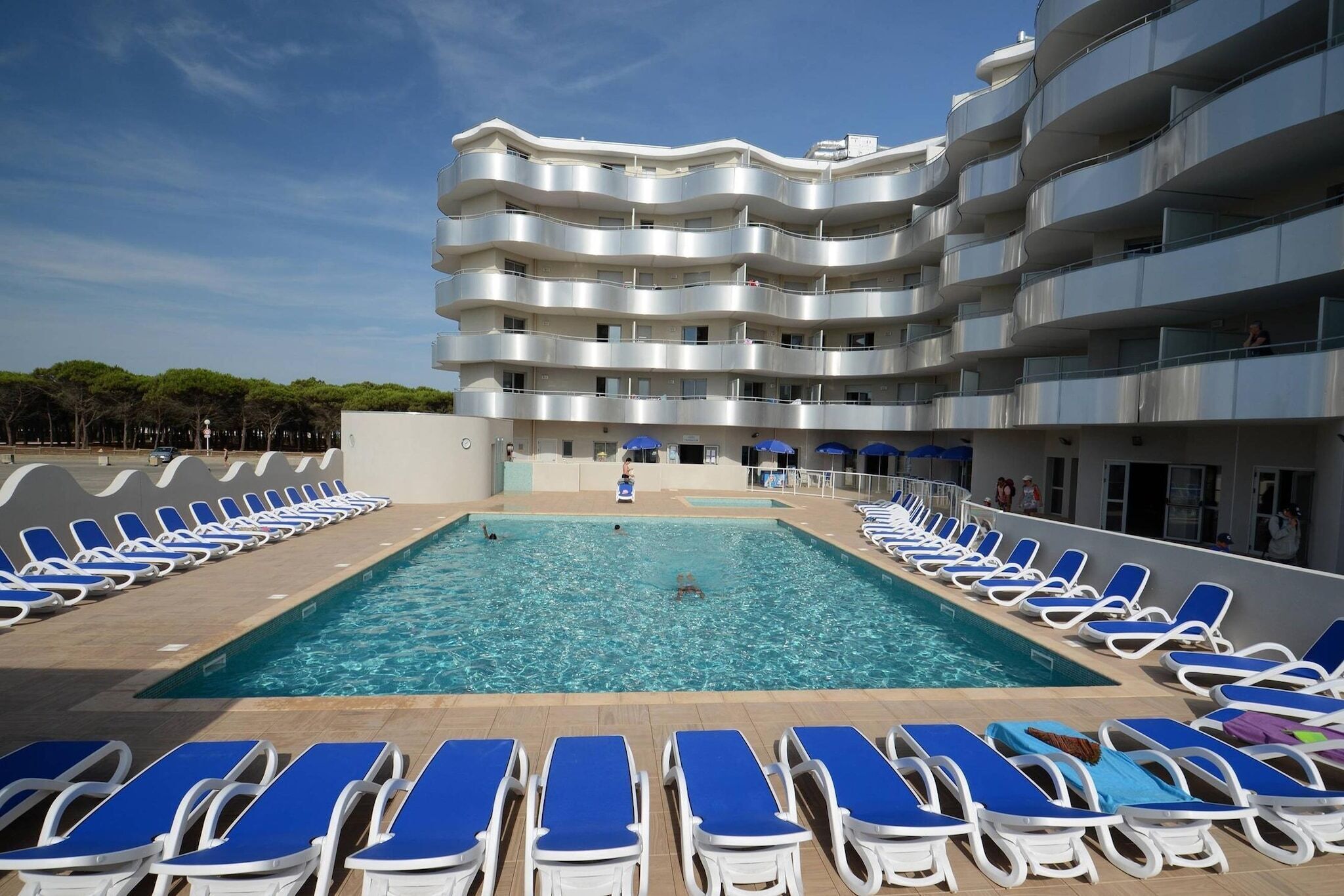 Une piscine avec des chaises longues et un immeuble à plusieurs étages doté de balcons.