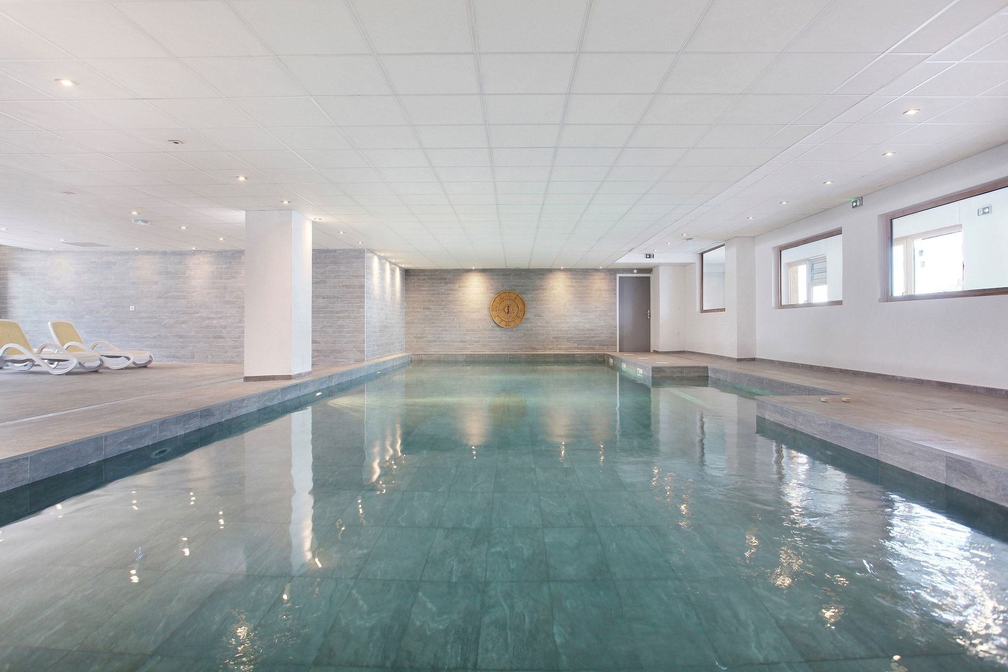Una amplia piscina interior con agua cristalina, rodeada de paredes con un diseño texturizado y un reloj en la pared.