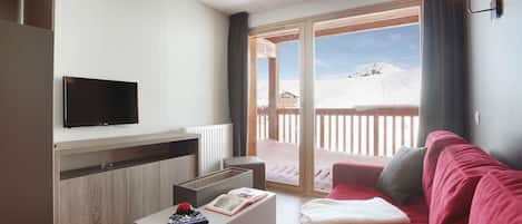 A modern living room with a flat-screen TV, a wooden cabinet, a red sofa, a white coffee table, and a balcony with a view of a snowy landscape.