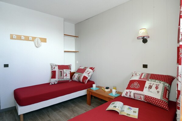 A room with two single beds, a small table with a book and a cup, and a wall-mounted light fixture.