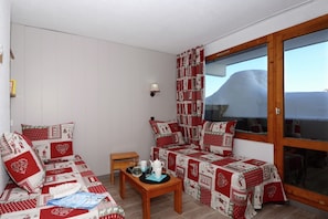 A cozy room with a sofa and a bed, both covered in red and white patterned blankets. There's a small wooden table with a kettle and glasses, and a window with curtains. - Apartment by Table Verte Lift (Les allues)