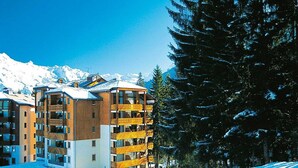 A multi-story hotel with balconies, surrounded by snow-covered mountains and pine trees.