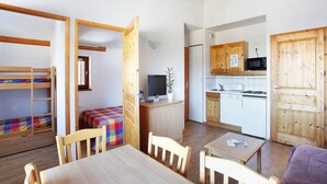 A compact room with a bunk bed, a small table, chairs, a television, a microwave, and a kitchen area with cabinets and a dishwasher.
