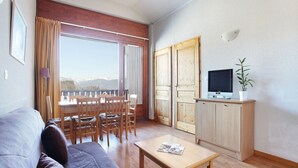 A living room with a sofa, a dining table with chairs, a television on a cabinet, and a view of mountains through a large window.